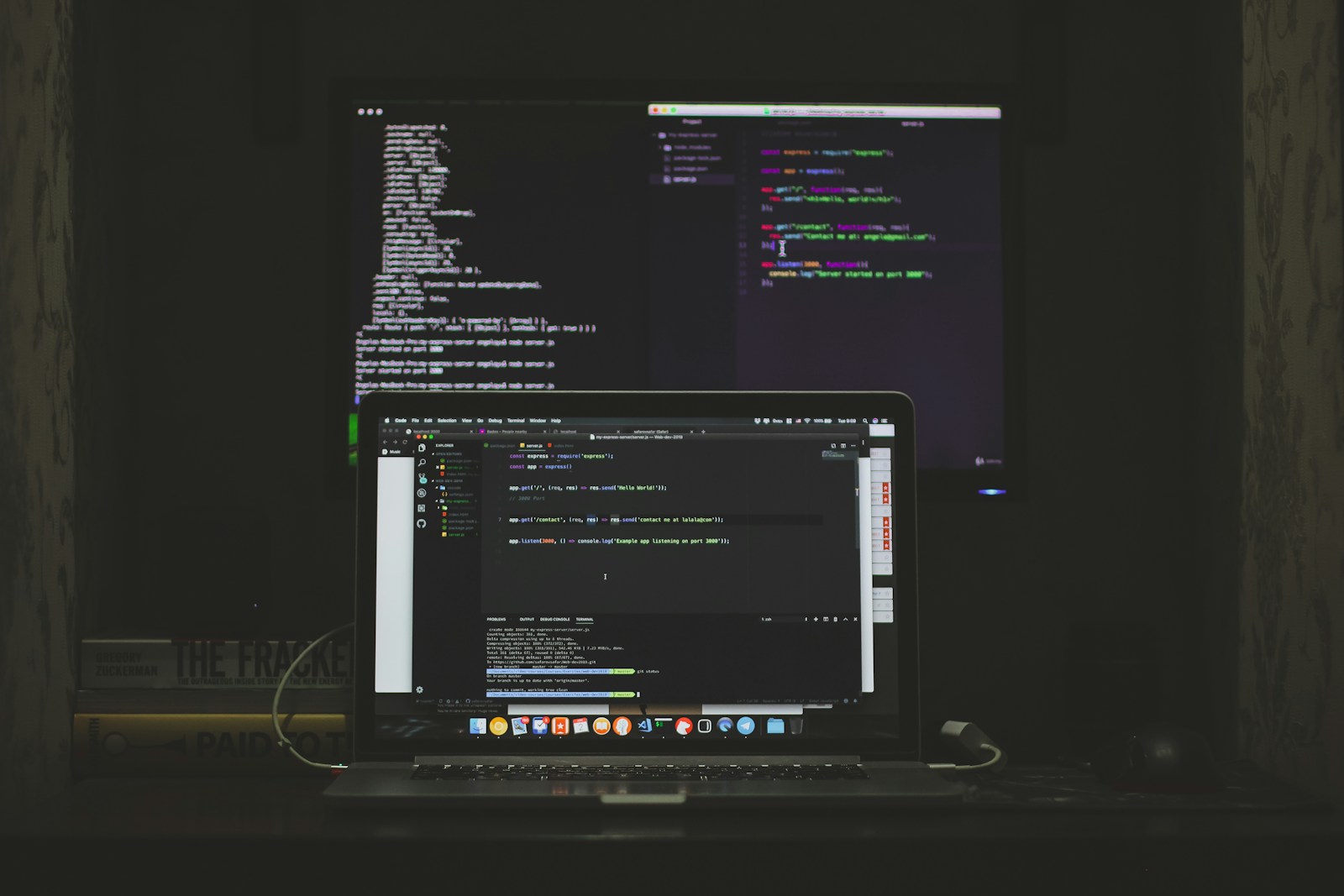 Dim-lit indie dev workspace: MacBook with VS Code open in front of a larger monitor full of source code